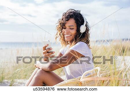 Stock Photograph - Young woman selecting music for headphones in beach dunes, Tuscany, Italy. Fotosearch