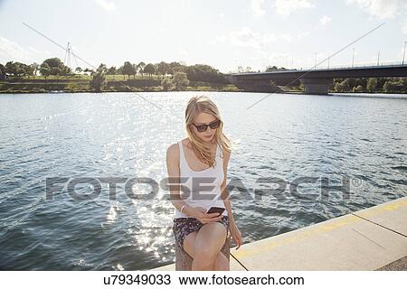 Young woman sitting at riverside reading texts on smartphone, Danube Island, Vienna, Austria View Large Photo Image Stock Image - Young woman sitting at riverside reading texts on smartphone, Danube Island, Vienna, Austria. Fotosearch