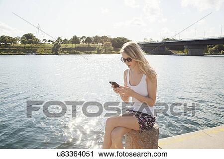 Young woman sitting at riverside texting on smartphone, Danube Island, Vienna, Austria View Large Photo Image Stock Image - Young woman sitting at riverside texting on smartphone, Danube Island, Vienna, Austria. Fotosearch