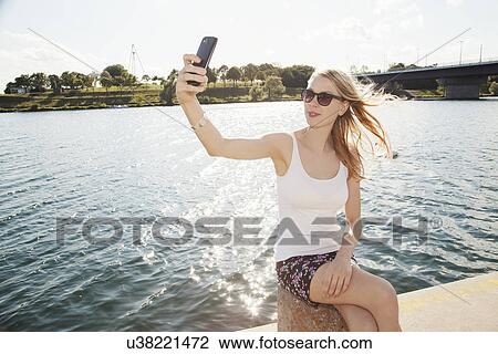 Stock Image - Young woman sitting at riverside taking smartphone selfie, Danube Island, Vienna, Austria. Fotosearch
