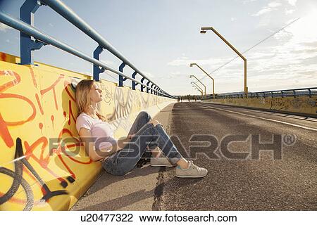Stock Image - Young woman sitting leaning against graffiti wall on bridge in sunlight. Fotosearch