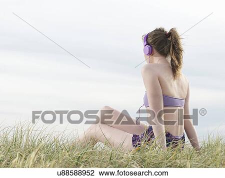 Stock Image - Young woman sitting on grass, wearing headphones. Fotosearch