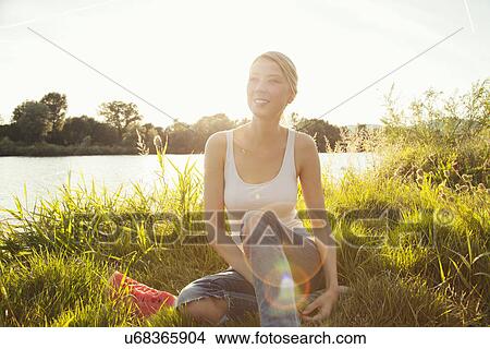 Picture - Young woman sitting on grassy riverbank, Danube Island, Vienna, Austria. Fotosearch