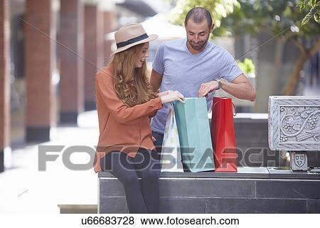Young woman sitting on wall with shopping bags, man looking in bags View Large Photo Image Stock Photo - Young woman sitting on wall with shopping bags, man looking in bags. Fotosearch