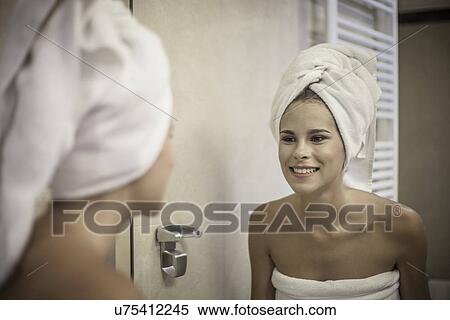 Stock Photography - Young woman wearing towel on head looking at reflection in mirror. Fotosearch