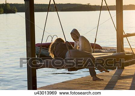 Stock Photo - Female friends enjoying on swing chair at dock, Lake of The Woods, Ontario, Canada. Fotosearch