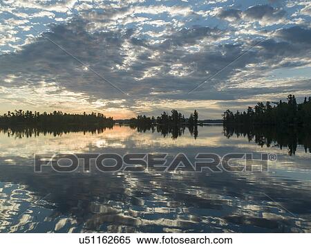 Stock Photography - Reflection of trees and clouds on water at dusk, Lake of The Woods, Ontario, Canada. Fotosearch