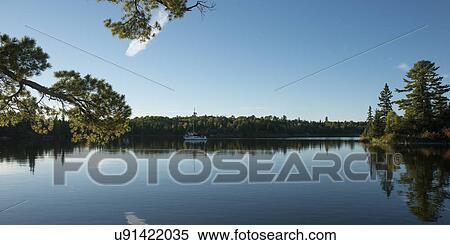 Stock Photography - Reflection of trees with a sail boat, Kenora, Lake of The Woods, Ontario, Canada. Fotosearch