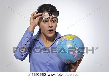 Stock Image - Amazed young businesswoman looking at globe over glasses against gray background. Fotosearch