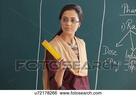 Stock Photograph - An angry young female teacher holding ruler against chalkboard . Fotosearch