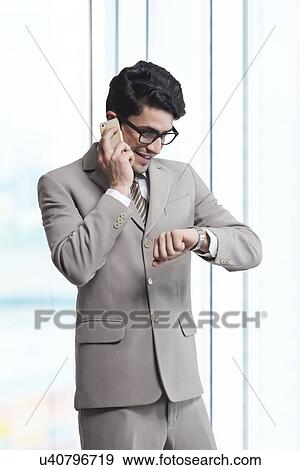 Businessman answering mobile phone while checking time in office View Large Photo Image Stock Photo - Businessman answering mobile phone while checking time in office. Fotosearch