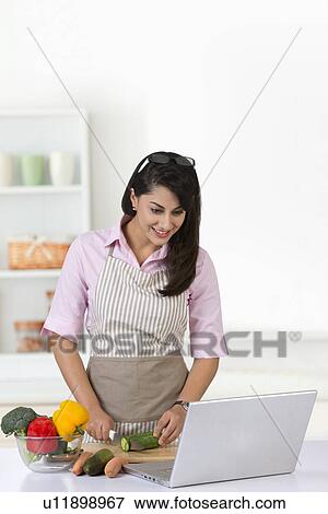 Stock Photo - Businesswoman using laptop while chopping vegetable in kitchen. Fotosearch