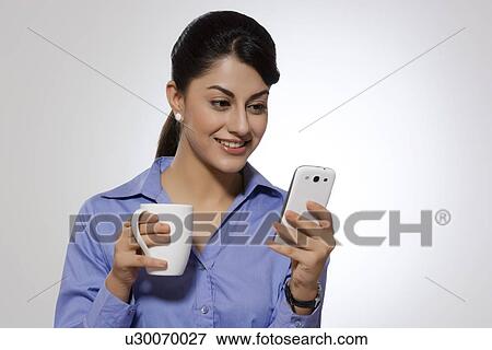 Stock Photo - Businesswoman with coffee mug reading text message on smart phone against gray background. Fotosearch