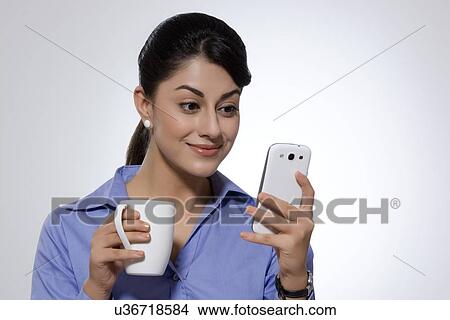 Picture - Businesswoman with coffee mug smiling while reading text message on smart phone over gray background. Fotosearch