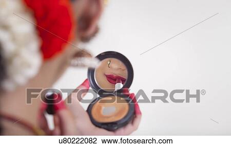 Stock Image - Close-up of Bharatanatyam dancer applying lipstick in compact mirror over white background. Fotosearch