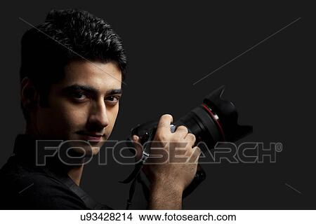 Close-up of young male photographer holding camera against black background View Large Photo Image Picture - Close-up of young male photographer holding camera against black background . Fotosearch