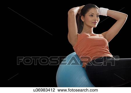 Stock Image - Fitness young woman balancing on exercise ball with hands behind head over black background . Fotosearch