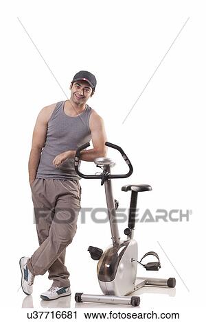 Stock Image - Full length of a young man standing next to an exercise bike over white background . Fotosearch