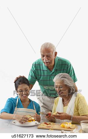 Stock Image - Grandmother and granddaughter working in the kitchen while grandfather looks on. Fotosearch