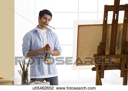 Stock Image - Handsome young artist with paintbrush in mouth standing in front of painting . Fotosearch