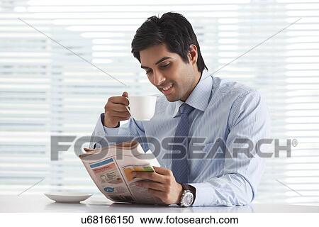 Stock Image - Handsome young businessman reading newspaper while having a cup of tea in office . Fotosearch