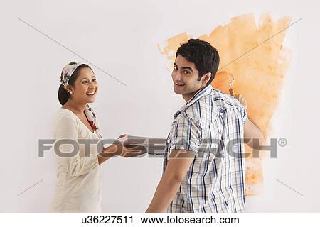 Stock Image - Handsome young man smiling with woman while applying paint on wall . Fotosearch