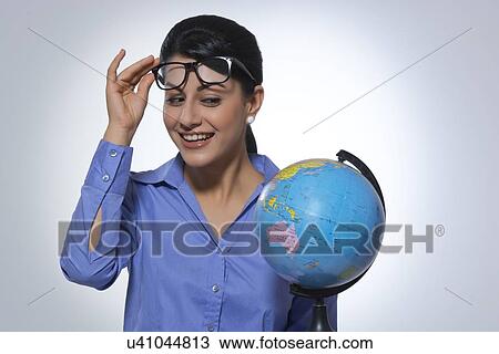 Stock Image - Happy businesswoman looking at globe over glasses against gray background. Fotosearch