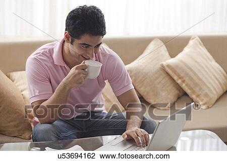 Happy man sipping coffee while using laptop at home View Large Photo Image Stock Image - Happy man sipping coffee while using laptop at home . Fotosearch