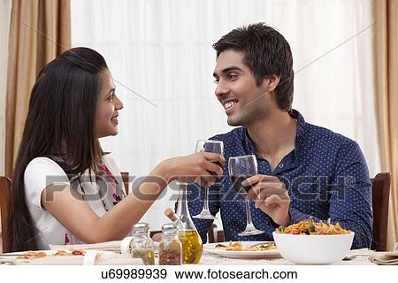 Stock Photo - Happy young couple clinking wine glass while looking at each other at restaurant . Fotosearch