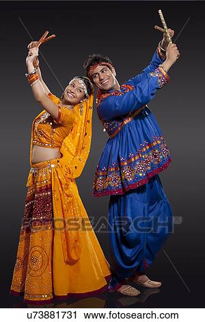 Stock Image - Happy young Indian couple performing Dandiya Raas over black background. Fotosearch
