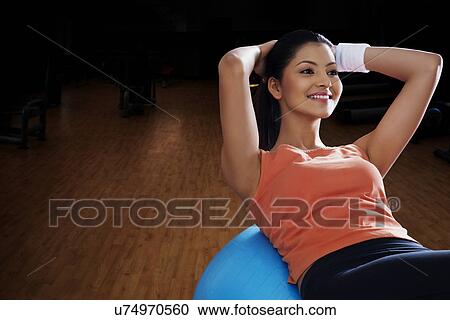 Stock Image - Happy young woman balancing on exercise ball with hands behind head . Fotosearch