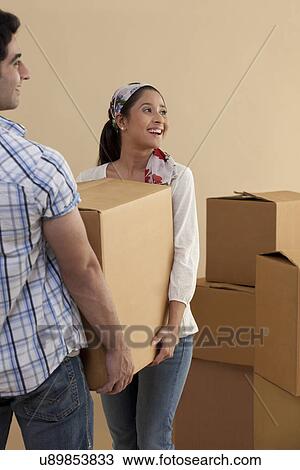 Stock Image - Happy young woman carrying cardboard box with man while moving in . Fotosearch