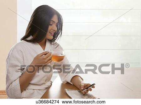 Picture - Happy young woman text messaging while holding tea cup at dining table . Fotosearch