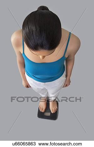 Stock Image - High angle view of young woman standing on weighing scale against gray background . Fotosearch