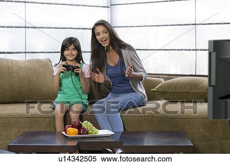 Stock Photography - Little girl playing video game with mother sitting besides her on sofa at home . Fotosearch