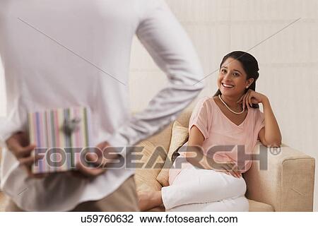 Stock Image - Man hiding surprise gift behind his back for woman at home . Fotosearch