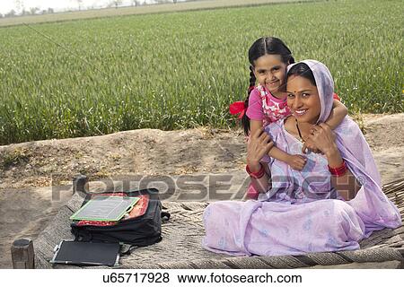 Stock Photo - Portrait of a happy Indian mother and daughter sitting on cot with field in background . Fotosearch