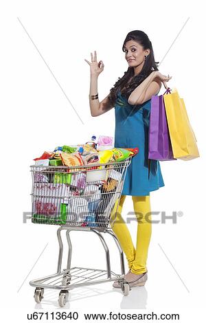 Stock Image - Portrait of a woman with a shopping cart and shopping bags giving ok sign. Fotosearch