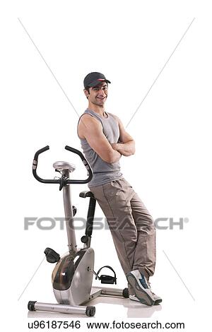 Stock Photograph - Portrait of a young man with arms crossed leaning on exercise bike over white background . Fotosearch
