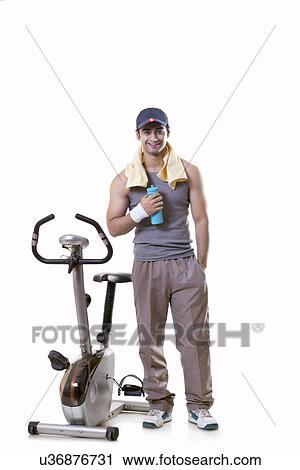 Stock Image - Portrait of a young man with water bottle standing next to an exercise bike over white background . Fotosearch