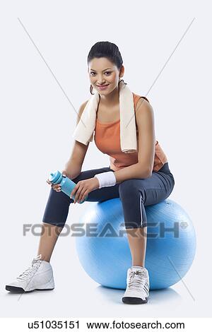 Stock Image - Portrait of a young woman with towel round shoulders sitting on a fitness ball over white background . Fotosearch