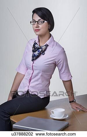 Stock Image - Portrait of confident businesswoman sitting on office desk. Fotosearch