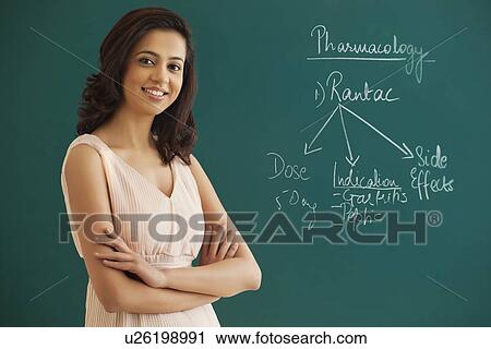Stock Image - Portrait of female teacher with arms crossed standing against green board . Fotosearch