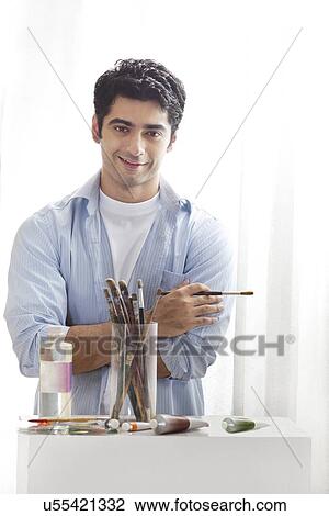 Stock Image - Portrait of handsome young man holding paintbrush in art studio . Fotosearch