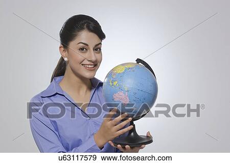 Stock Photo - Portrait of happy Indian businesswoman holding globe against gray background. Fotosearch