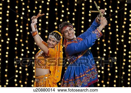 Picture - Portrait of happy Indian couple in traditional wear performing Dandiya Raas against neon lights. Fotosearch