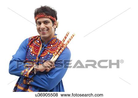 Stock Photo - Portrait of happy young man in traditional wear holding dandiya over white background. Fotosearch