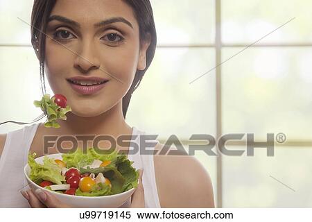 Stock Photography - Portrait of happy young woman eating salad of lettuce, cherry tomatoes and mushrooms against glass window . Fotosearch