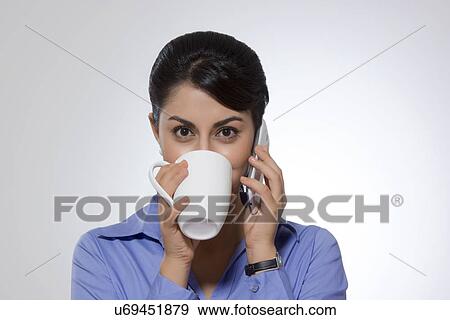 Stock Photo - Portrait of Indian businesswoman drinking coffee while answering phone against gray background. Fotosearch