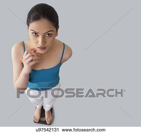 Portrait of worried young woman standing on weighing scale against gray background View Large Photo Image Stock Image - Portrait of worried young woman standing on weighing scale against gray background . Fotosearch